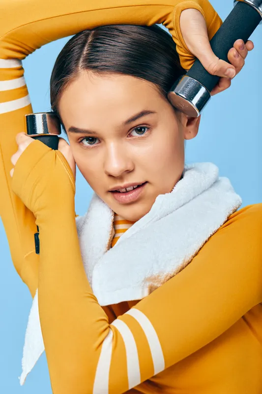 Athletic woman in orange sportswear lifts dumbbells, towel around her neck, mid motion in a bright studio with a cool blue background, showcasing fitness determination and strength.