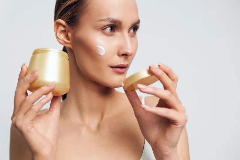 A young woman applies cream on her cheek while holding an open jar of skincare product in a bright studio setting. Concept for beauty routine, hydration, and facial care.
