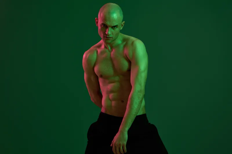 Athletic man posing in low light with an intense expression. The striking green backdrop enhances the mood and emphasizes his well-defined physique.