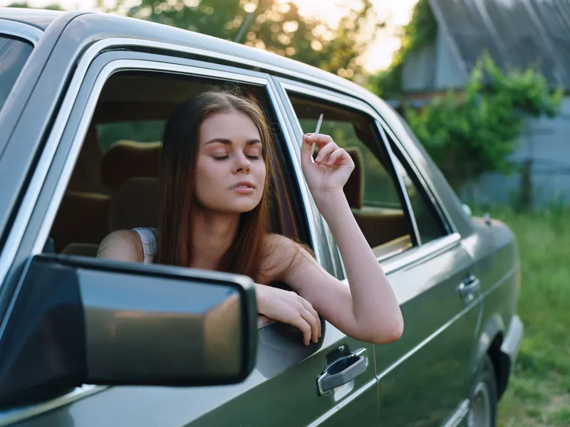 Young woman smoking a cigarette in a vintage car, serene expression, sunlit outdoor background, showcasing a blend of nostalgia and tranquility.