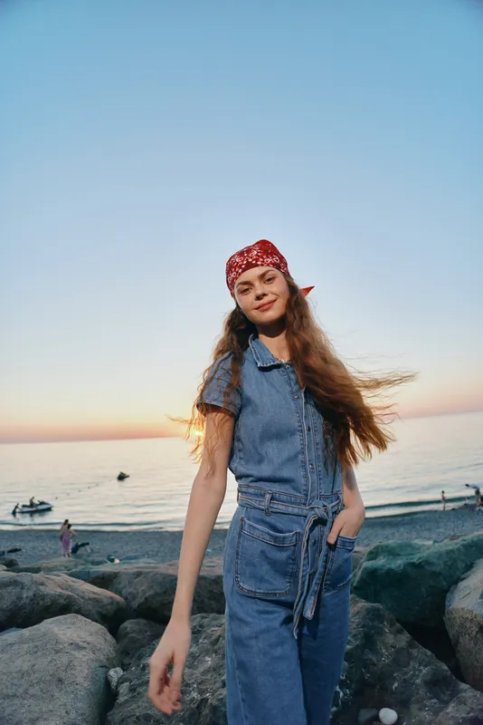 lifestyle woman in modern boho-western denim street style with red bandana by the sea at sunset, natural light and film color tones creating relaxed vibe
