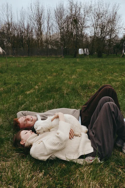 Lifestyle couple cuddling on the grass in sweaters, a goat kid nearby, a warm moment in the countryside. Soft natural light creates a peaceful atmosphere, intimate connection, and cozy, relaxed mood