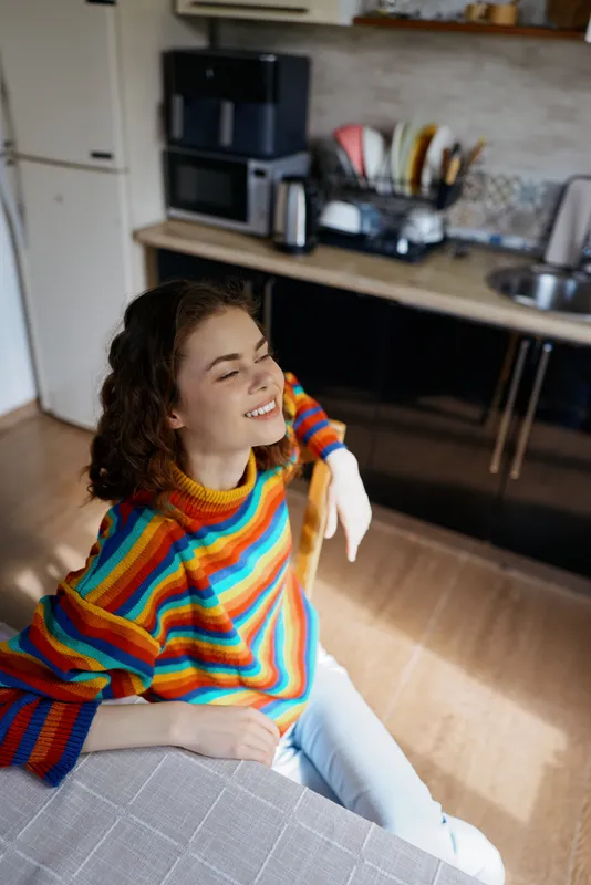 Cheerful woman indoors at home, wearing a colorful striped sweater, smiling in a bright kitchen, relaxed casual pose, warm sunlight, cozy atmosphere and natural colors across the scene