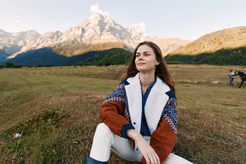 Woman portrait sitting in meadow with mountain landscape, enjoying nature and travel outdoors. Casual jacket, thoughtful expression and scenic field for hiking and relaxation.