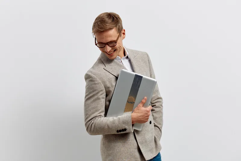 Confident young man wearing glasses and a light grey blazer holding a silver laptop in his hands, smiling happily, looking down, isolated on a plain light-colored background, professional business sty