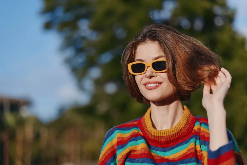 Woman smiling in colorful striped sweater and yellow sunglasses, short bob hair and casual outfit, touching hair outdoors in sunny park with warm sunlight, lifestyle travel and fashion portrait.