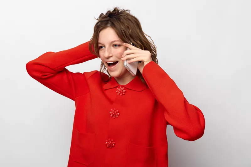 Young woman in red sweater talks on smartphone with excited expression, isolated on white background. Concept of communication, happiness, and casual lifestyle.