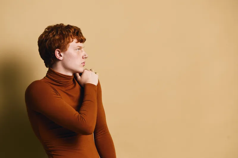 Profile portrait of young man with red hair wearing brown turtleneck sweater, looking thoughtful against beige background. Casual style with natural light and contemplative mood.