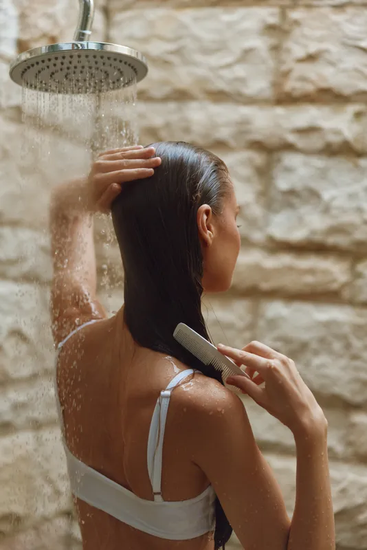 woman combing long wet hair under outdoor shower with stone wall background, natural light, skincare and hygiene routine, tranquil moment