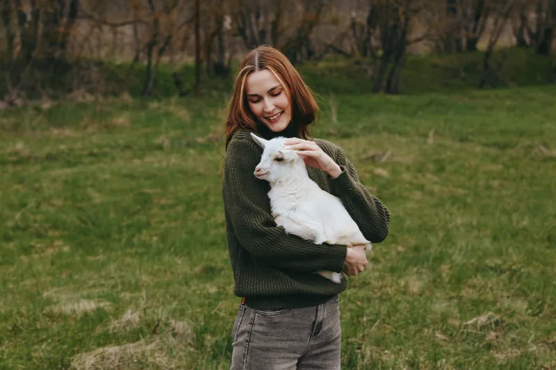 A woman holds a goat kid in a grassy field, outdoors, showcasing lifestyle, affection, cuddle, nature through warm, natural light and a calm countryside mood.