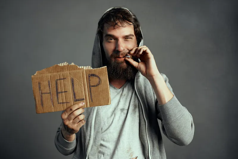 Бородатый мужчина в капюшоне держит картонный знак с надписью HELP