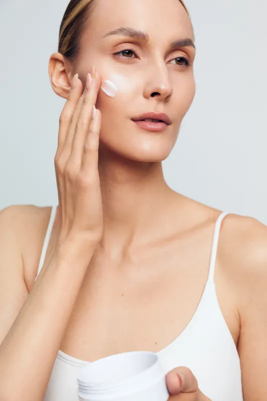 Young woman applying face cream with fingers, clear skin, natural makeup, white tank top, close-up portrait, skincare routine, soft lighting, neutral background
