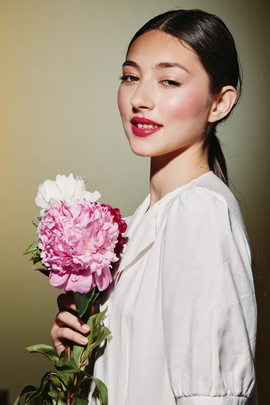 Young woman with glossy hair holds pink peony flowers while wearing soft makeup and a white blouse. Studio lighting highlights her fresh radiant skin. Concept for beauty, fashion, floral.