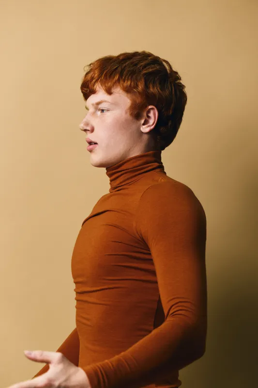 Young man with red hair wearing a brown turtleneck shirt posing sideways on beige background. Casual style with expressive facial profile and modern look for fashion or portrait.