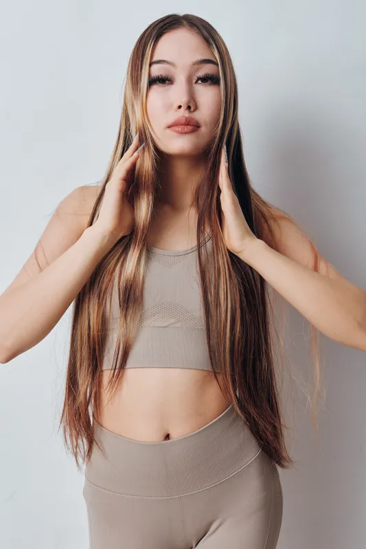Young woman with long straight hair wearing beige crop top and leggings, posing with hands near face on white background, beauty portrait with natural makeup and calm expression.