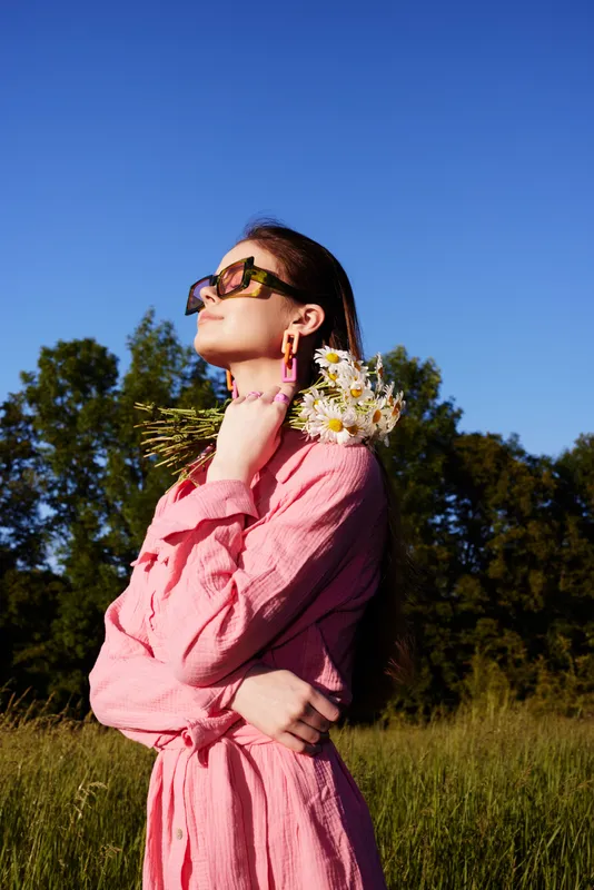 Young woman in pink dress holding flowers, enjoying sunny day outdoors, expressing joy and serenity against blue sky and green backdrop