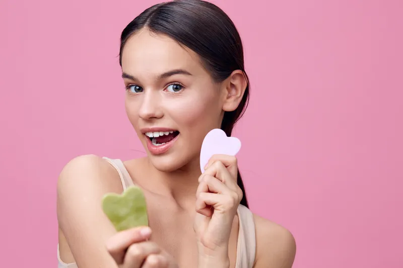 Young woman holding heart shaped beauty tools on a pink background, expressing joy and confidence, showcasing skincare concepts and modern beauty trends