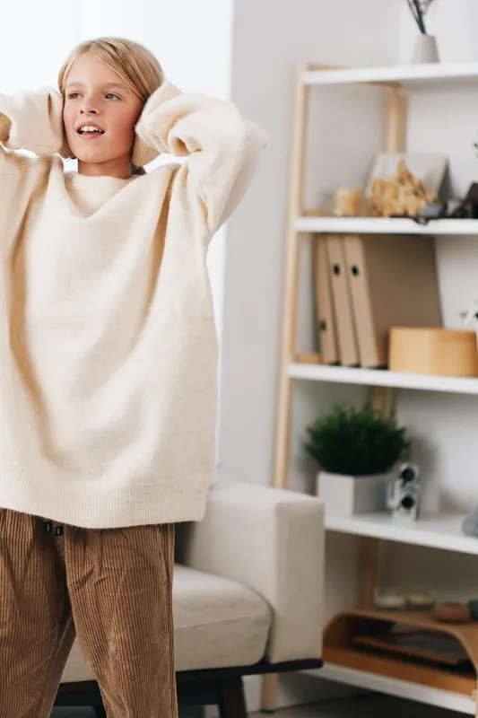Young child stands indoors wearing a soft cream sweater and corduroy pants, hands raised to adjust hair, exuding a cozy, relaxed mood in a warm, minimalist home setting.