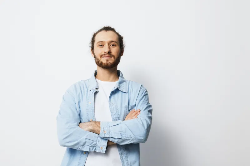 Young man with beard posing confidently, wearing casual denim shirt on a white background, showcasing positivity and self assurance