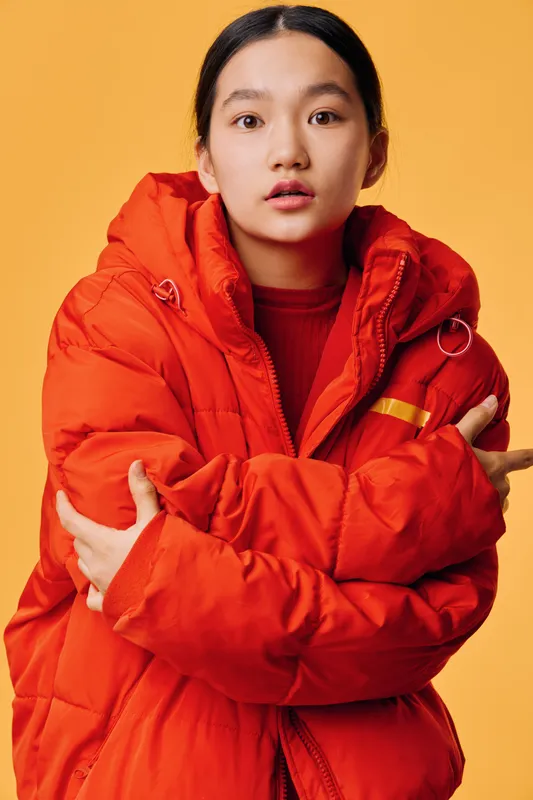 Young woman in bright orange winter jacket with arms crossed looking at camera against yellow background, casual fashion portrait for cold season style and outerwear concept.