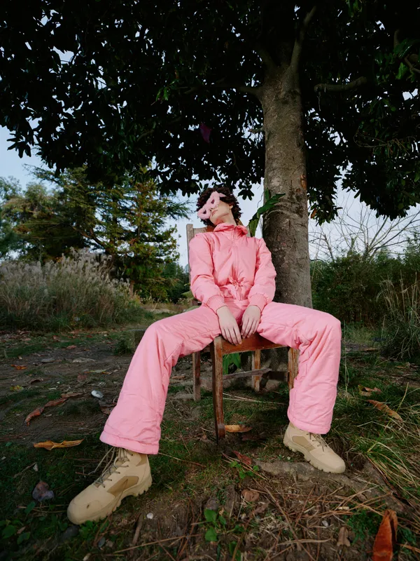 Outdoor fashion scene with a person in a pink suit sitting beneath a large tree, legs stretched, boots on ground, relaxed pose in a natural park setting, conveying casual style and calm confidence.