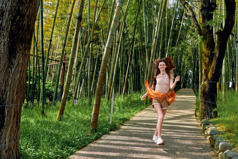 Woman girl running on a bamboo forest path, smiling in a dress and skirt with sneakers. Happy young woman enjoying nature on a sunny trail, playful motion, freedom and summer leisure outdoors.