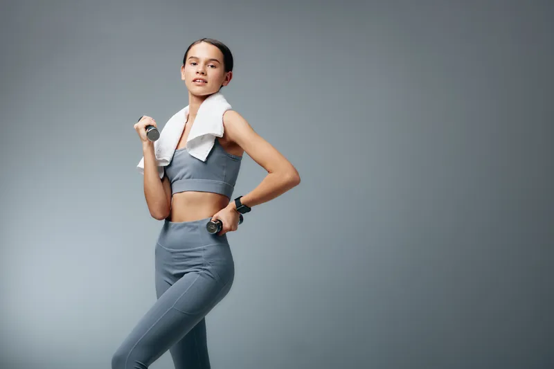 Female fitness model in athletic wear with a dumbbell, towel draped over shoulder, mid workout in a sleek studio. Focus on strength, balance, and dedication during training.