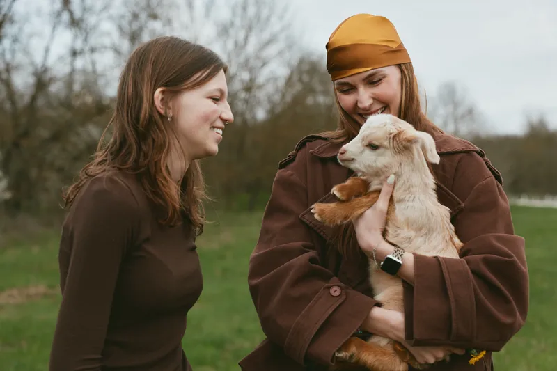 Two young women enjoy a casual moment outdoors as one holds a baby goat while they smile and share a warm connection in a natural lifestyle setting.