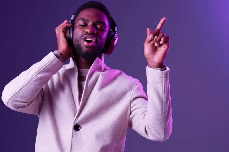 Happy young Black man with headphones dancing to music in stylish beige coat against vibrant purple background, expressing joy and emotion