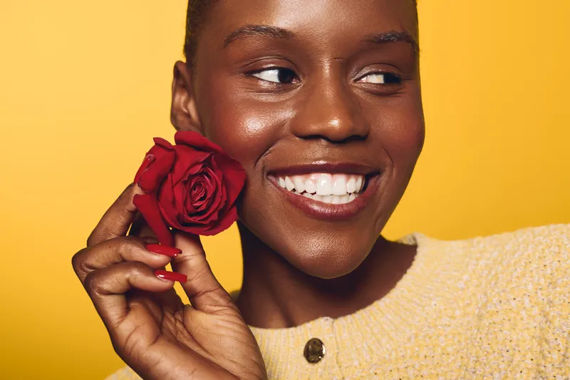 Smiling dark skinned African woman holding red rose near face on yellow background. Concept of beauty, happiness, floral, natural skin, and positive emotions in portrait photography.