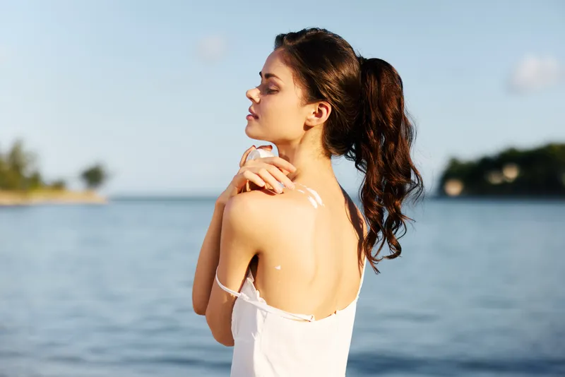 A young woman stands by the sea, bare shoulders, white dress, sunlight on skin, windswept hair, and a serene expression, capturing quiet summer coastal beauty.