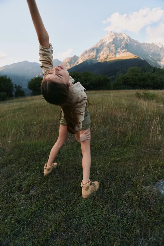 Joyful woman outdoors performing a handstand in a grassy field, mountains in the background, sunlight highlighting a carefree, active lifestyle and natural colors, bright.