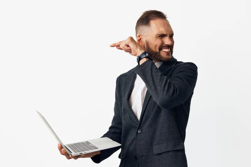 Professional man in a suit stands against a plain studio backdrop, expressing confidence and energy while using a laptop. His determined expression conveys ambition and focus in a corporate setting.