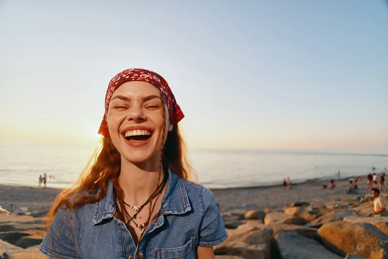 smiling woman in modern boho-western denim street style with film color effect, wearing red bandana and layered necklaces by the beach at sunset