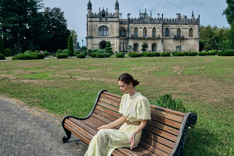 Woman in a light dress sits on a wooden bench in a tranquil park with a historic castle in the background, conveying calm, elegance, and contemplative mood