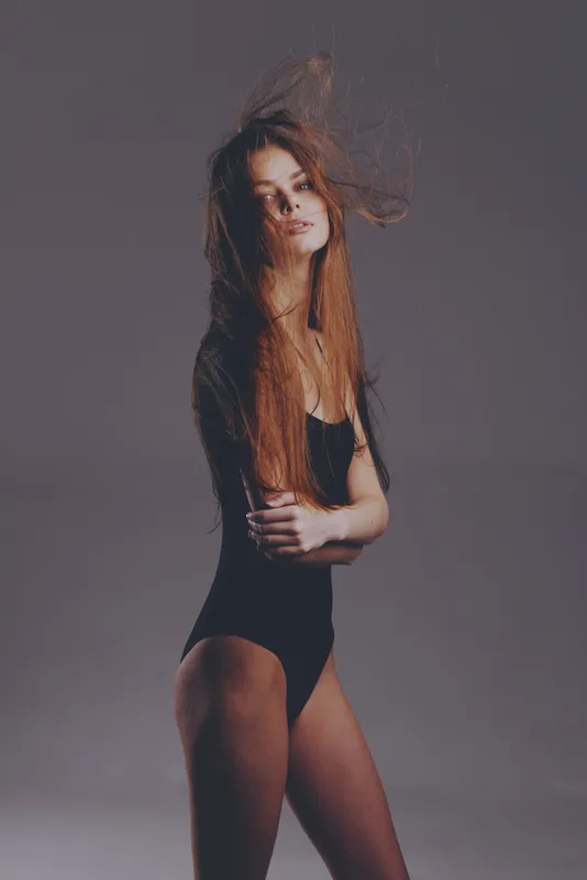 slender woman with long red hair posing in black bodysuit against grey background