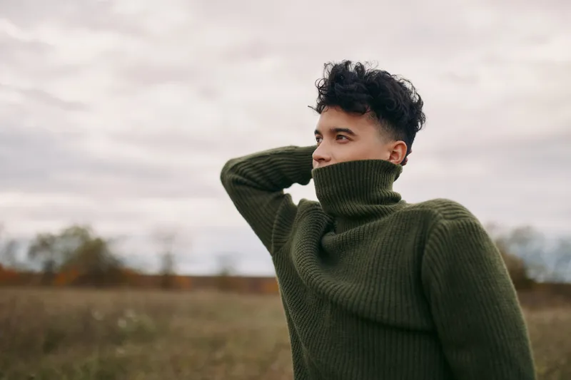 Young man in a green turtleneck sweater stands contemplative in an open field, face partly hidden, conveying authentic presence and genuine emotion with subtle natural expression.