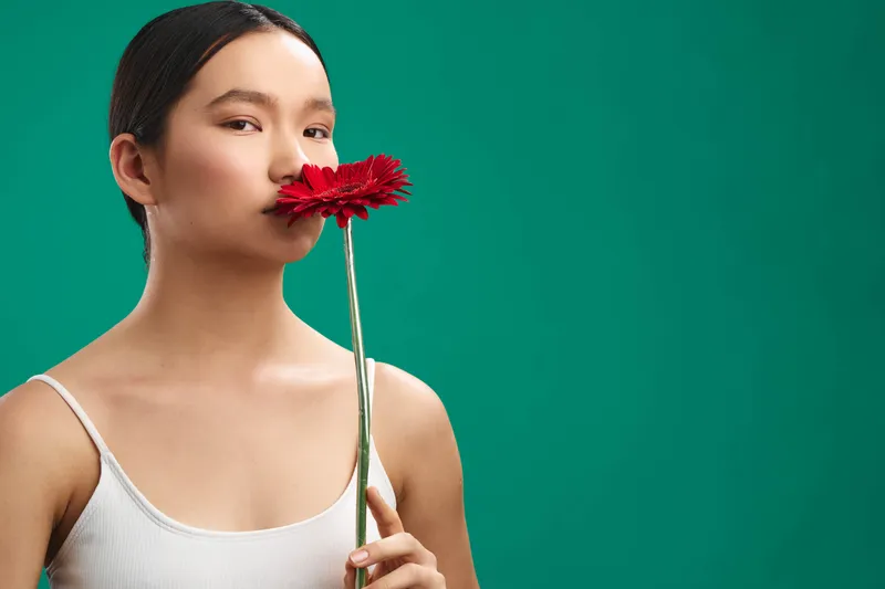 Asian woman stands against a solid green background, gently holding a red flower near her lips. She gazes softly forward, conveying calmness, poise, and quiet elegance.