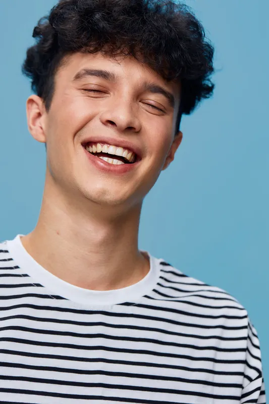 Smiling man in striped shirt laughing joyfully against a solid blue background, expressing happiness and carefree positive emotion in casual attire and curly hair.
