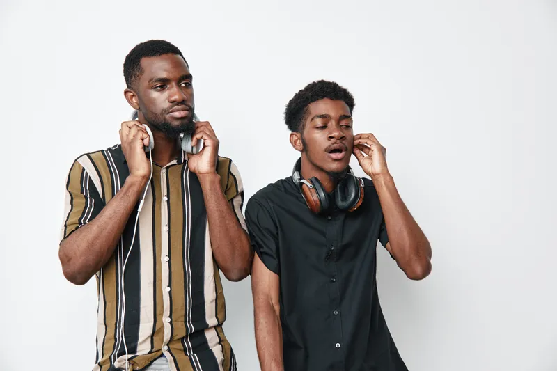 Two african american men with dark skin wearing headphones standing against white background, one with striped shirt and the other in black shirt listening to music.