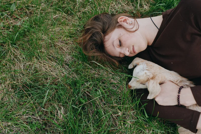 A calm moment outdoors with a young woman resting on grass while cradling a small goat kid, showcasing gentle bond, nature, and comfort in a peaceful lifestyle scene.