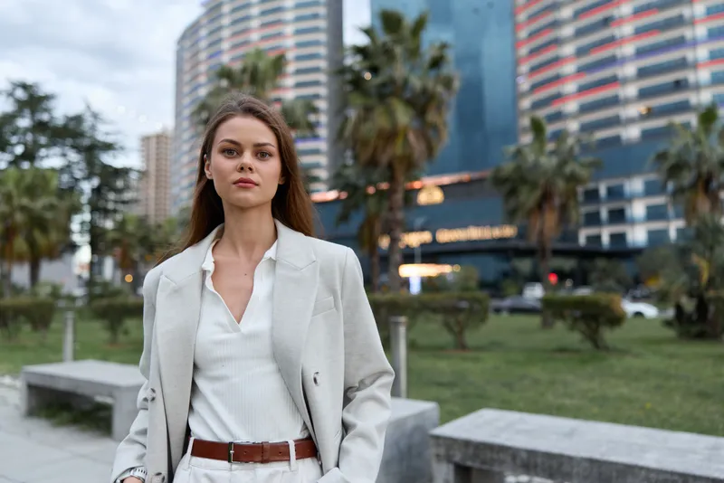 Confident woman in business attire walking outdoors in urban setting with modern buildings and palm trees, showcasing professional style and determination