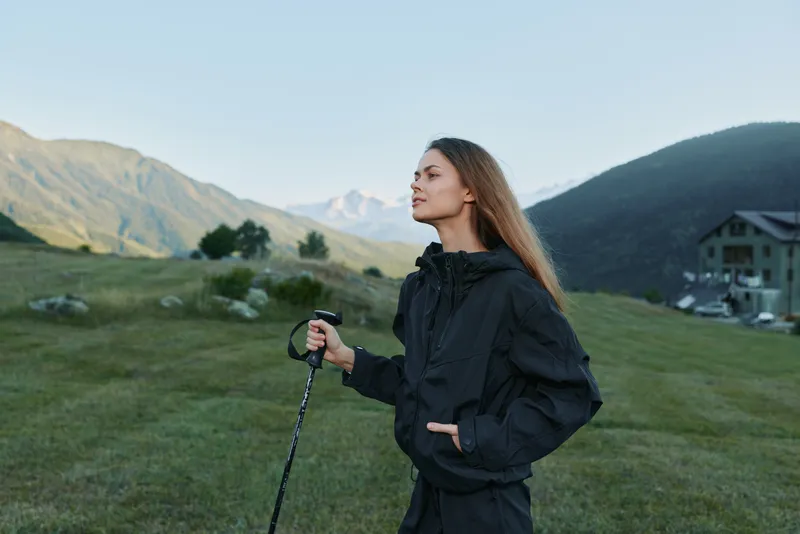 A focused hiker woman stands in a grassy mountain landscape, holding a trekking pole. She wears a dark jacket and gazes toward distant peaks, exuding adventure and resilience.