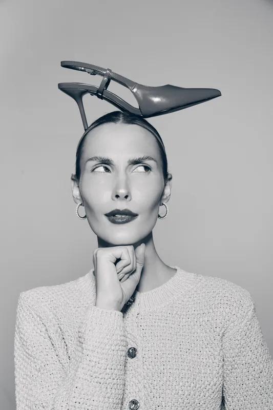 Young Caucasian woman with light skin wearing a knitted sweater balances a high heel shoe on her head, looking thoughtful on grey background.