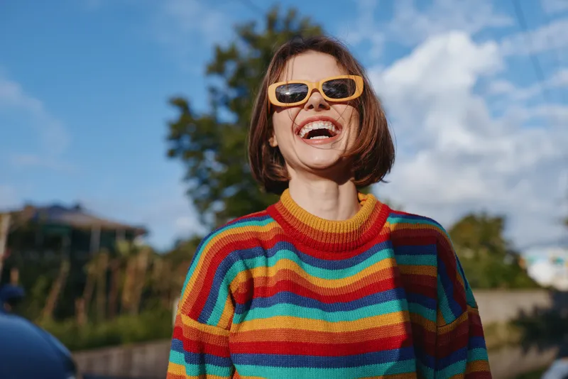Woman smiling and laughing in a colorful striped sweater and yellow sunglasses, short hair bob, casual outfit while standing outdoors in sunny suburban street for lifestyle travel.