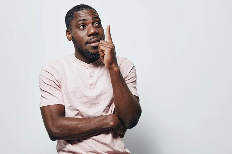 African American man with dark skin wearing light pink shirt standing with crossed arms and finger raised, thinking or having an idea on plain white background in studio.