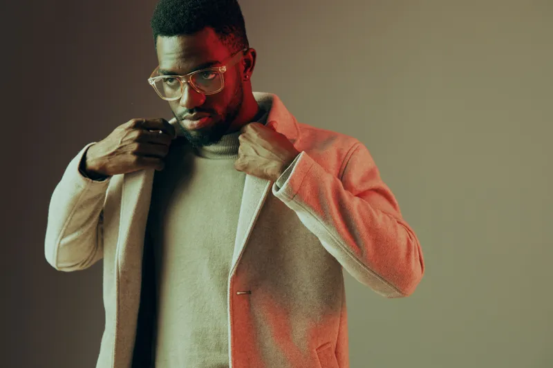 A fashionable Black man adjusting his stylish coat in a minimalist studio with soft pastel tones, showcasing modern elegance and confidence