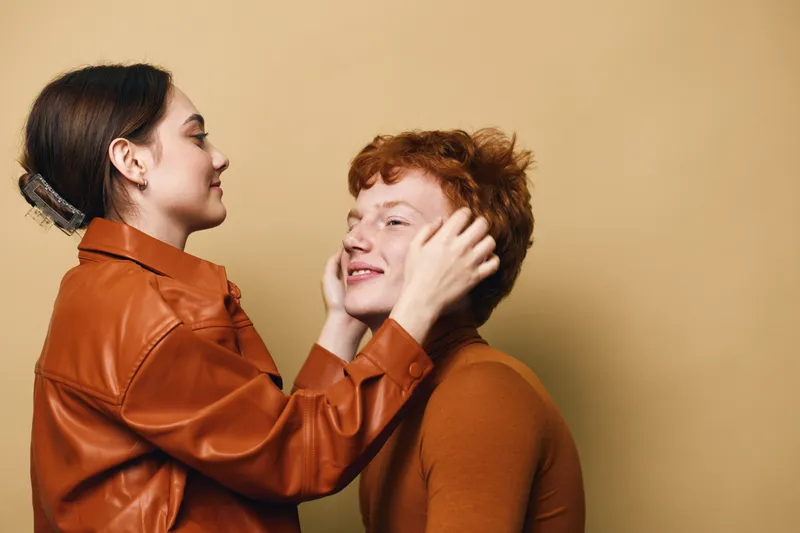 Young couple touching faces and looking at each other with gentle smile, wearing brown and orange clothing with plain beige background in studio.