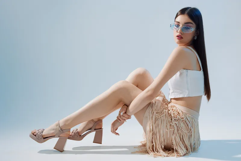 Young woman beauty fashion model wearing sunglasses and stylish fringe skirt sits on floor against light background. Concept for summer style, elegance, and confident attitude.