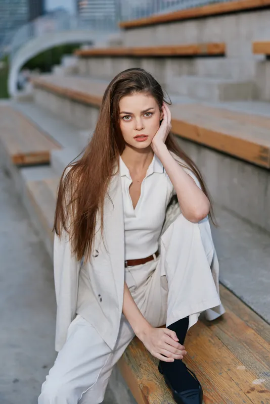 Stylish woman in white suit sitting on wooden city steps with thoughtful expression, urban fashion portrait with natural light and casual elegance